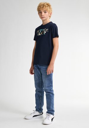 Marineblauwe katoenen T-shirt met een kleurrijke print met het woord "REBEL." Gecombineerd met lichtblauwe jeans en witte sneakers met zwarte accenten.