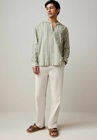 Gestreiftes Leinenhemd in Grün und Weiß, leicht und mit Kragen. Kombiniert mit cremefarbener Leinenhose und braunen Riemchensandalen.
