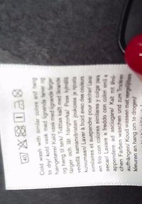 Clothing care label with washing symbols and multilingual instructions held by a hand with red-painted nails against a dark background.