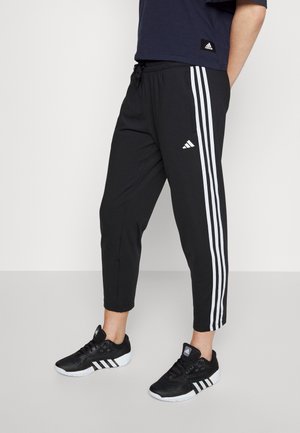 Person iført sorte Adidas træningsbukser med hvide striber kombineret med sorte Adidas sneakers på en ensfarvet baggrund.