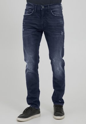 Dunkelblaue Denim-Jeans in Slim Fit, mit subtilen Used-Effekten, kontrastierenden Nähten und Fronttaschen. Kombiniert mit schwarzen Sneakers.