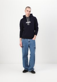 Marinblå hoodie med krämfärgade snören och framficka, i kombination med ljusblå, oversized cargo-jeans och svarta skor. Enkelt, avslappnat design.