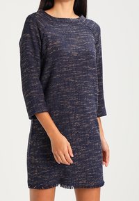 Modelka w teksturowanej sukience w granatowym kolorze z rękawami o długości trzech czwartych i postrzępionym dołem, pozująca na neutralnym tle.