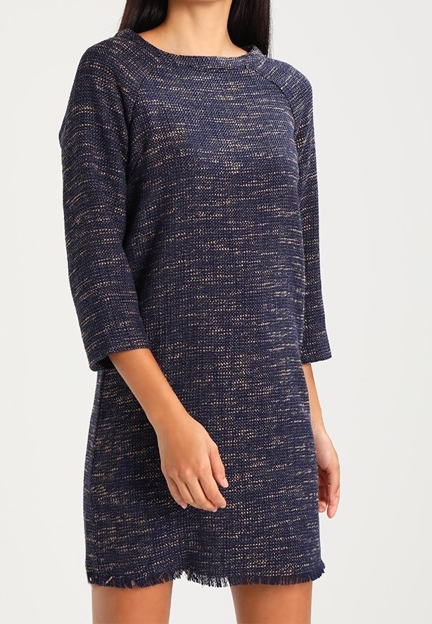 Modelka w teksturowanej sukience w granatowym kolorze z rękawami o długości trzech czwartych i postrzępionym dołem, pozująca na neutralnym tle.