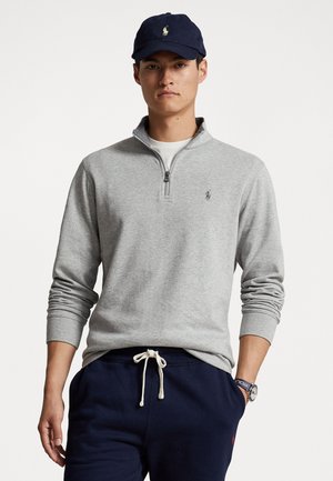 Gray zip-up sweatshirt with long sleeves, featuring a small logo on the chest and paired with dark navy sweatpants and a navy cap.