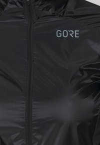 Czarna, lekka kurtka z pełnym zamkiem z przodu, logo GORE, gładką teksturą i dopasowanym krojem, idealna do aktywności na świeżym powietrzu.