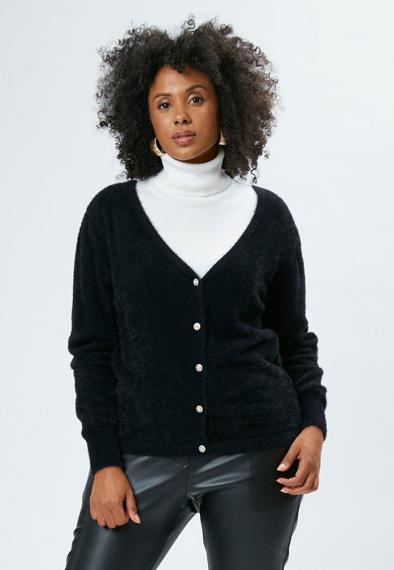 Schwarzer, flauschiger Cardigan mit V-Ausschnitt und Knopfverschluss, verziert mit perlenähnlichen Akzenten. Trägt man über einem weißen Rollkragenpullover, kombiniert mit schwarzen Hosen.