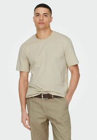Beige Kurzarm-T-Shirt mit Rundhalsausschnitt, aus weichem Stoff. Getragen mit hellbraunen Hosen und einem passenden Gürtel.