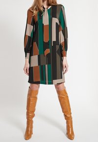 Robe à manches longues avec un motif géométrique en vert, marron et beige. Tissu lisse avec un nœud au niveau du col et coupe décontractée.