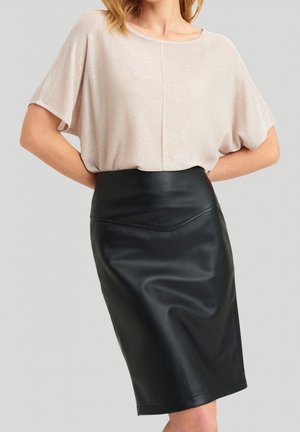 Mujer con una camiseta beige de manga corta y una falda lápiz negra de cuero de talle alto, de pie con las manos detrás de la espalda.