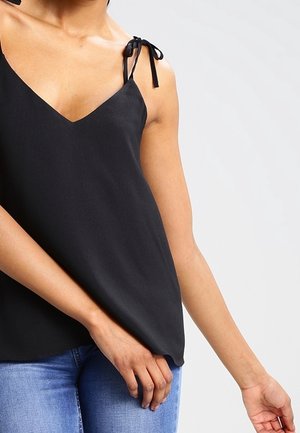 Femme portant un haut noir sans manches avec des bretelles à nouer et un jean bleu, visible des épaules aux hanches sur un fond uni.