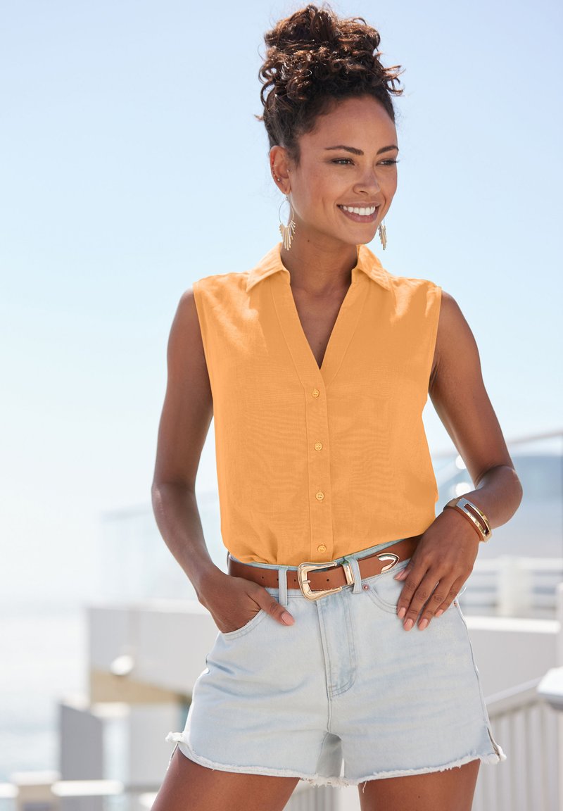 Sleeveless, collared shirt in light orange fabric, with a button front. Worn with light blue denim shorts and a brown belt.