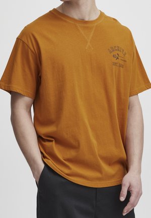 Person iført en orange kortærmet t-shirt med "ARCHIVE EST. 1999" og en rose grafik på brystet, hænder i lommerne på sorte bukser.