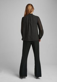 Blusa nera trasparente con motivo sottile e scollatura arricciata, abbinata a pantaloni a pince neri a vita alta e tacchi.