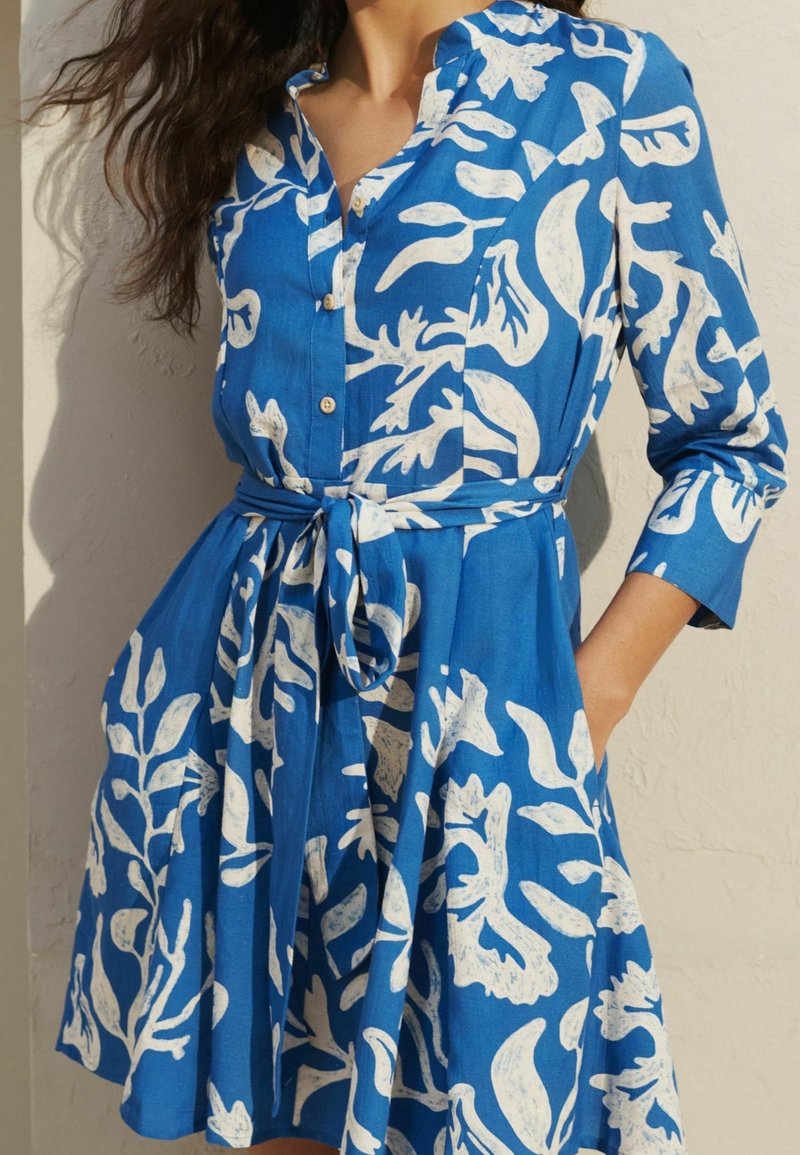 Femme portant une robe bleue boutonnée avec des motifs botaniques blancs et une ceinture à la taille, debout avec une main dans la poche contre un mur clair.