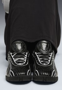 Scarpe da ginnastica nere con accenti lucidi e lacci bianchi. Supporto alla caviglia imbottito distintivo e design testurizzato, posizionato su un indumento scuro.
