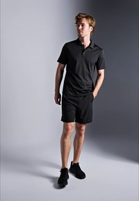 Jovem a caminhar vestido com uma camisola polo preta, calções pretos e ténis pretos contra um fundo degradé cinza.