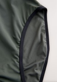 Tela verde oscuro con ribete elástico negro, probablemente parte de una prenda, mostrada sobre un fondo blanco liso.