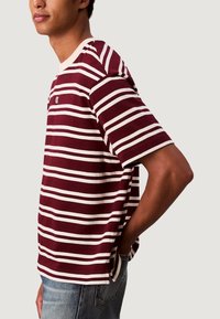 Červeno-krémové pruhované tričko s krátkým rukávem, kulatým výstřihem a postranními rozparky. Látka vypadá měkce a má volný střih. Viditelné malé logo.