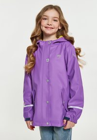 Lila vattentät jacka med huva, silverfärgade tryckknappar, reflexdetaljer och ett gult foder, bärd över en blå denim-outfit.