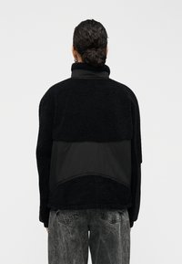 Μαύρο μπουφάν από φλίς με ψηλό γιακ ά και υφή υφάσματος. Διαθέτει λείο μαύρο πάνελ στη μέση και μαύρο τζιν με ξεθωριασμένη υφή.