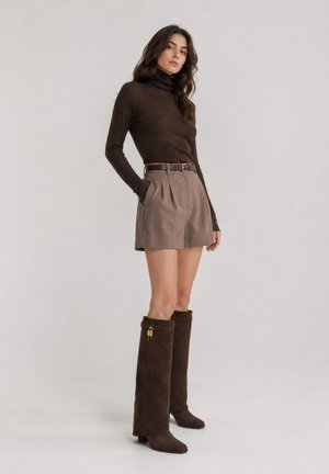 Mujer con un jersey de cuello alto marrón oscuro, pantalones cortos taupe de cintura alta con cinturón y botas altas de ante marrón oscuro con detalle de cierre dorado.