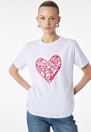 Joven con el cabello peinado hacia atrás lleva una camiseta blanca con arte lineal floral en forma de corazón rojo y texto "Romance", combinada con jeans azules.