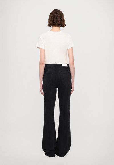 Jean noir à pattes larges avec texture rayée verticale, mettant en avant une taille haute et des poches arrière. Associé à un haut à manches courtes de couleur claire.