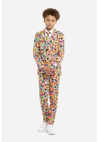 Complete con un abito a motivi colorati caratterizzato da pois multicolori su uno sfondo luminoso, abbinato a una camicia bianca e una cravatta coordinata, sneakers bianche.