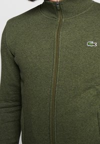 Zelená zipová bunda vyrobená z měkké tkaniny se stojáčkovým límcem. Obsahuje malé vyšití loga na hrudi a hladký zip.