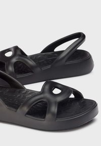 Sandales en caoutchouc noires avec un design à bout ouvert, larges brides croisées avec des découpes circulaires, semelle intérieure texturée et semelle à talon bas.