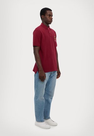 Burgunderfarget polo-skjorte med korte ermer og krage, med et brodert design på venstre bryst. Kombinert med lyseblå jeans og hvite sneakers.