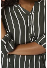 Blouse rayée verte avec des lignes verticales blanches, un col et des boutons. Manches longues retroussées avec un détail de poignet. Texture de tissu douce.