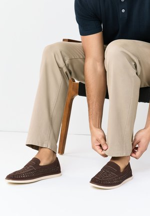 Homme assis sur une chaise en bois ajustant le bas d’un pantalon beige, portant des mocassins sombres en cuir tissé sur un sol blanc.