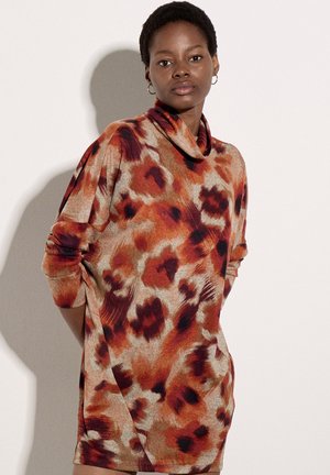 Femme aux cheveux courts portant un haut à manches longues et col montant avec un motif abstrait orange, marron et beige, debout contre un mur uni.