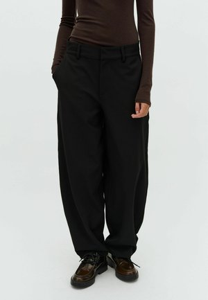 Personne portant un pantalon noir ample, un haut à manches longues marron foncé et des chaussures marron foncé brillantes, debout les mains dans les poches.