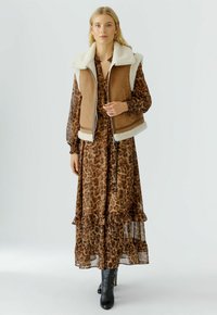 Frau in einem langen Kleid mit braunem Leopardenmuster, beiger Wildlederweste mit weißen Lammfellfutter und schwarzen Absatzstiefeln, die vor einem schlichten Hintergrund steht.