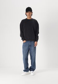 Črn oversized pulover, modri ravni jeans in beli športni čevlji, oblečeni s črno kapo; minimalistična zasnova z sproščenim krojem.