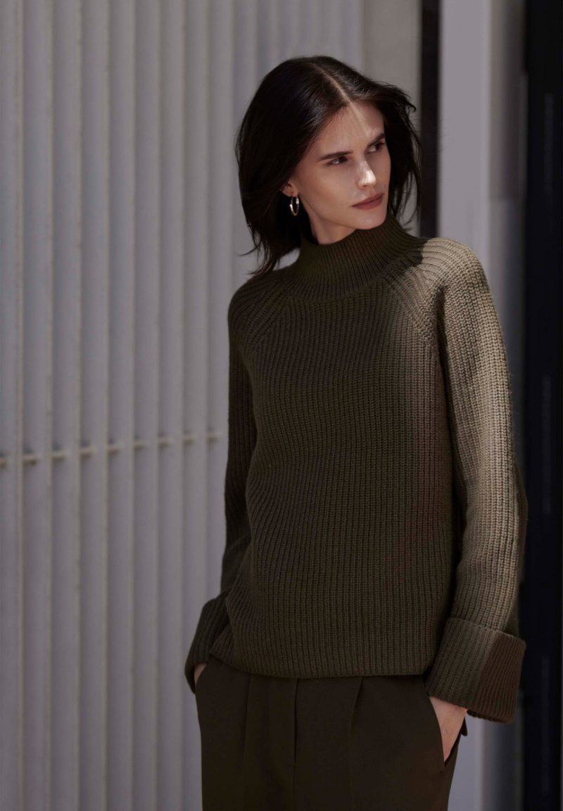Zielonooliwkowy sweter z dzianiny z wysokim kołnierzem, prążkowaną strukturą i szerokimi mankietami, noszony z ciemnymi spodniami na minimalistycznym tle.