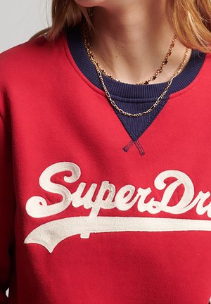 Sweat-shirt rouge arborant un logo "SuperDry" blanc, avec un col côtelé bleu marine et des chaînes dorées superposées autour du cou, mettant en valeur des textures variées.