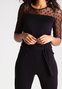 Zwarte top met stippen, transparante mouwen van gaas, aansluitend ontwerp en een gladde textuur. Heeft een ontspannen pasvorm en een taillebanddetail.