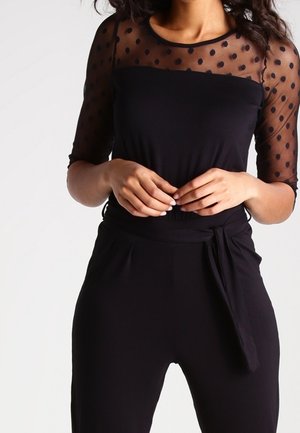 Black top with polka-dot translucent mesh sleeves, fitted design, and a smooth texture. Features a relaxed fit and waist tie detail.