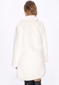 Donna che indossa un cappotto in pelliccia sintetica bianco fin sopra il ginocchio e stivali neri al ginocchio, vista da dietro su uno sfondo bianco semplice.