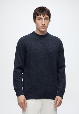 Junger Mann mit kurzen dunklen Haaren, der einen dunkelblauen Strickpullover und weiße Hose trägt, steht vor einem schlichten weißen Hintergrund.