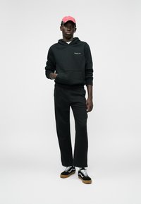 Sweatshirt preto com um bolso frontal, combinado com calças largas pretas e sapatilhas pretas com detalhes brancos e solas em borracha. Boné rosa com padrão.