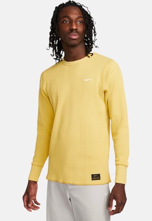 Gul, ribbet langærmet skjorte med rund hals, hvid Nike-logo på brystet og et sort mærke nær sømmen. Glat tekstur.
