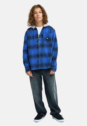 Jeune personne aux longs cheveux bouclés portant une chemise à carreaux bleue sur un t-shirt blanc, un jean sombre ample et des baskets noires, debout devant un fond uni.