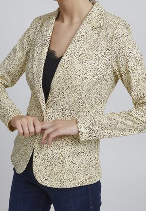 Mujer abrochándose un botón de una chaqueta amarilla con estampado de leopardo, sobre una blusa negra de encaje y vaqueros azul oscuro, contra un fondo gris claro.