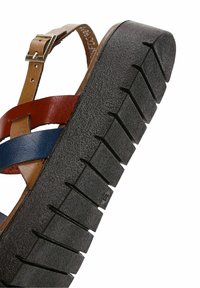 Sandal de cuero marrón y azul con una gruesa suela negra texturizada, que presenta un diseño de correas entrecruzadas y hebilla metálica ajustable.