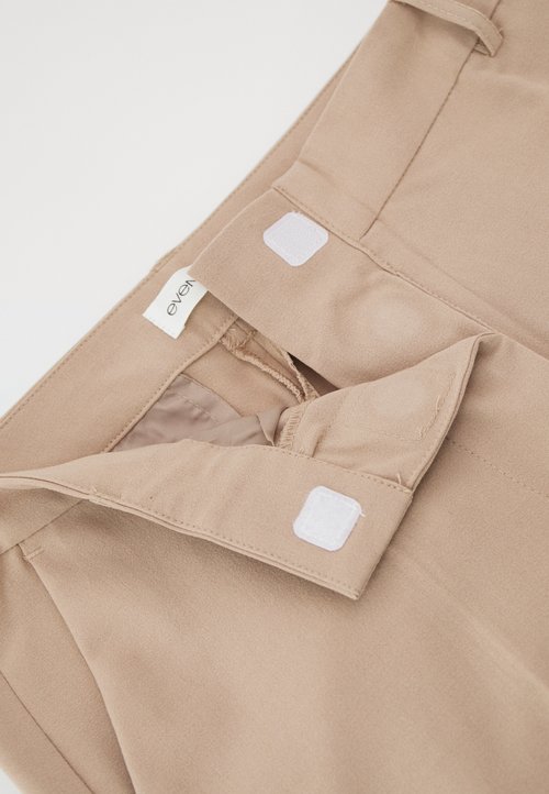 Top de tela beige con cuello, que presenta cierres de Velcro blancos y una pequeña etiqueta. Textura suave, líneas limpias y sin patrones.